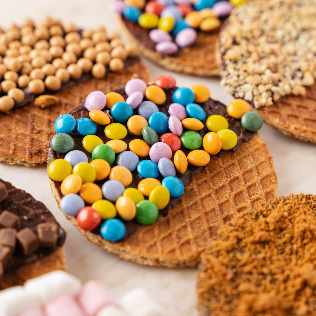 Close-up of fresh stroopwafels topped with chocolate, coloured candy-coated chocolates, caramel pearls and chopped nuts